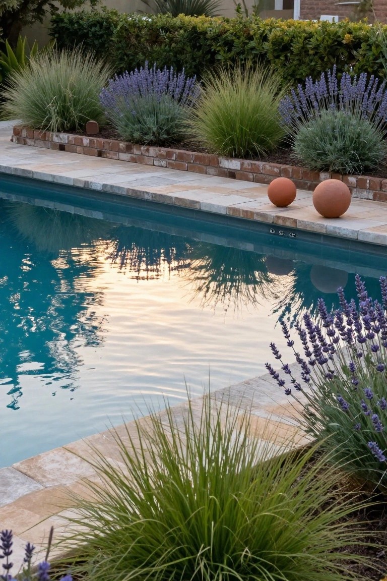 Swimming pool with beige travertine decking edged by raised brick planters containing ornamental grasses, lavender bushes, and silver-leaf plants, plus two orange terracotta spheres on the deck near lush green hedges in the background.