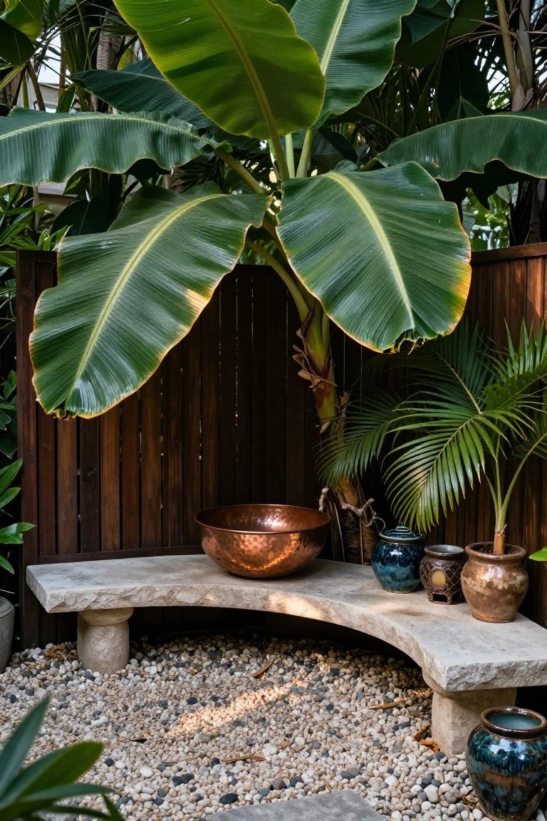 Curved stone bench with a large copper bowl on it in a fenced tropical garden area surrounded by banana plants, palms, pottery pots, and pebble ground cover.