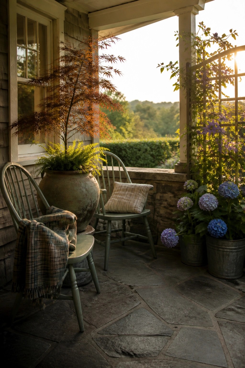 A covered stone porch with green wooden chairs, a large weathered pot holding a small tree and fern, smaller metal buckets with blue and purple hydrangeas, and a plaid blanket draped over one chair.