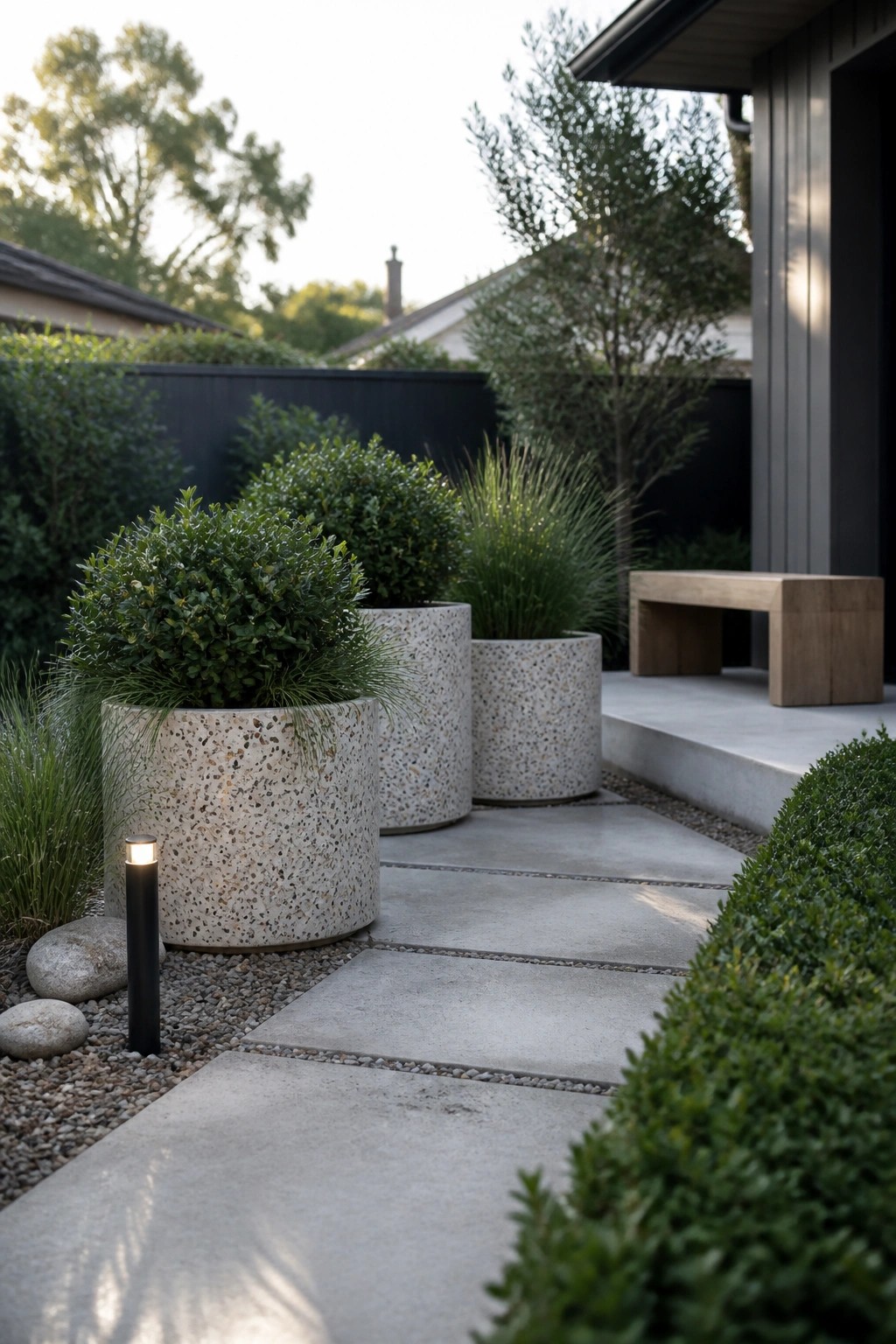 Modern garden with speckled concrete planters, pathway, wooden bench, and lit bollard.